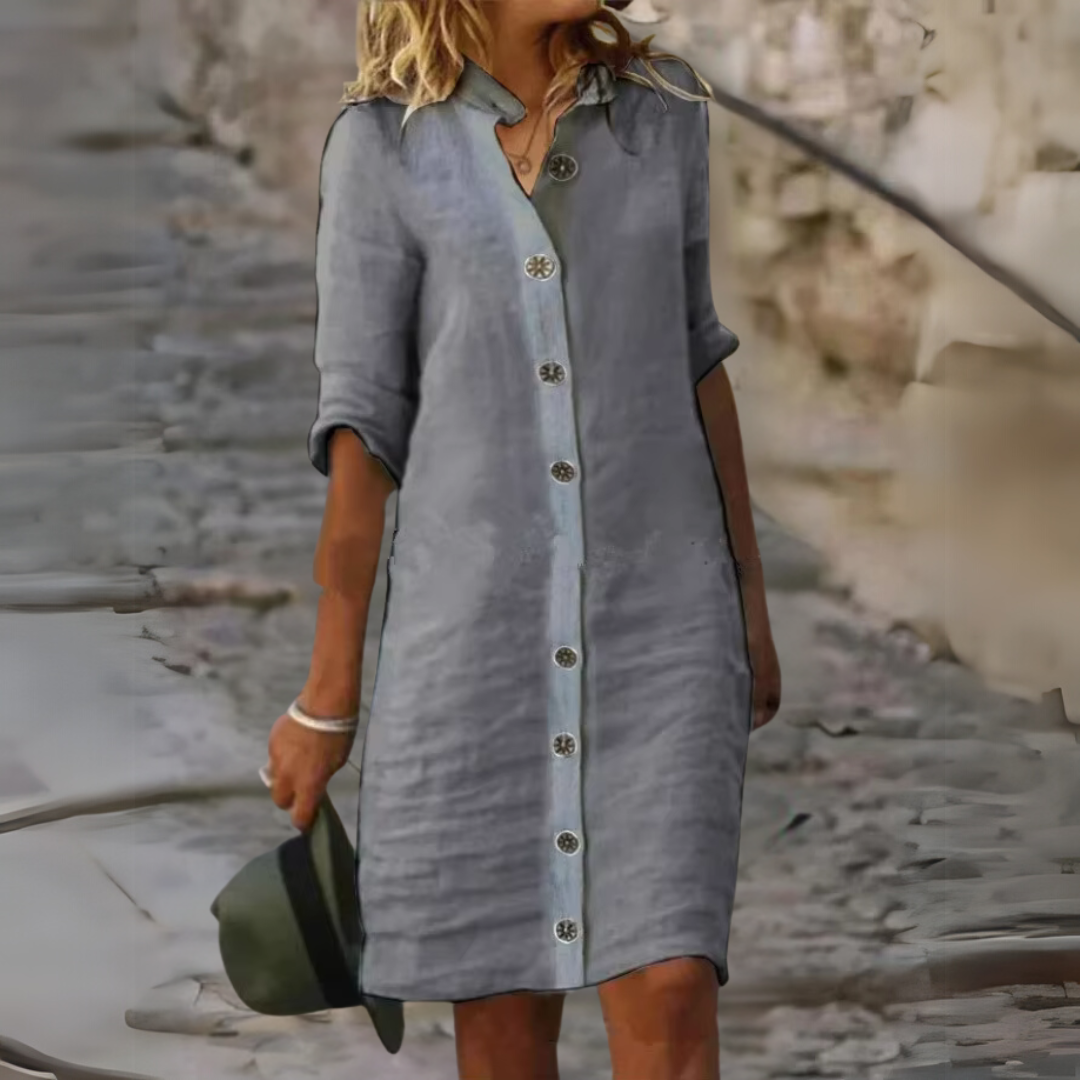 Frau in grauem Leinenkleid mit Knopfleiste, hält grünen Hut. Sommermode, lässiger Stil, modisches Outfit für Damen, urbaner Hintergrund.