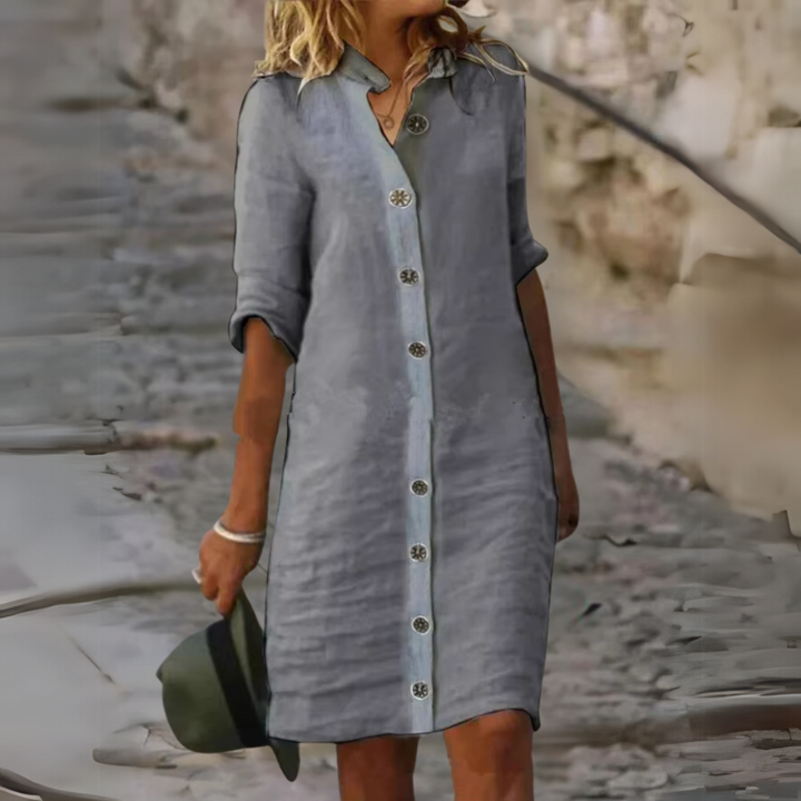 Frau in grauem Leinenkleid mit Knopfleiste, hält grünen Hut. Sommermode, lässiger Stil, modisches Outfit für Damen, urbaner Hintergrund.