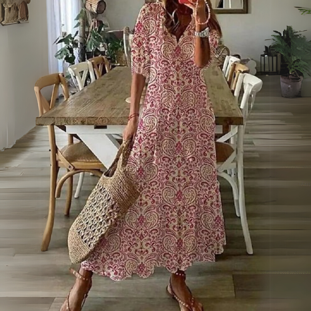 Frau in langem, geblümtem Sommerkleid mit Strohtasche steht in modernem Esszimmer. Holztisch, weiße Stühle, Pflanzen im Hintergrund.