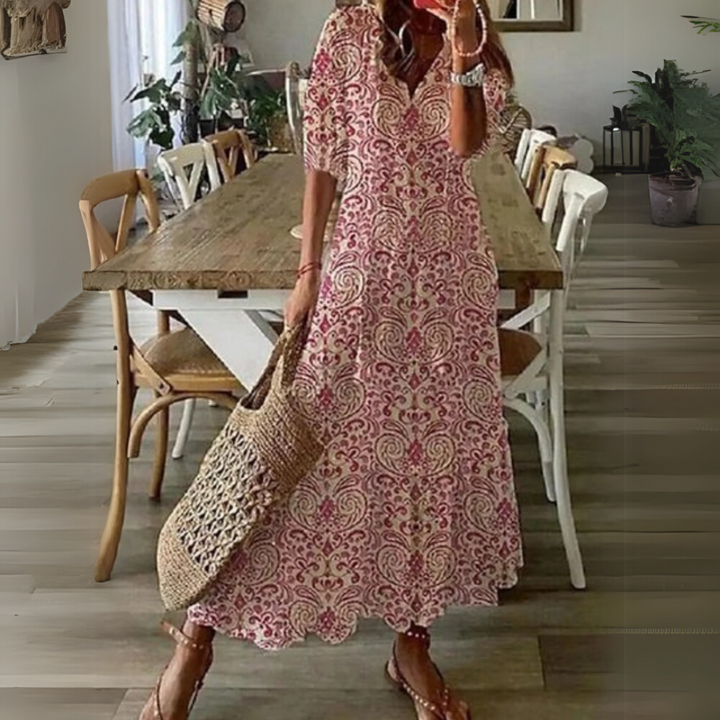 Frau in langem, geblümtem Sommerkleid mit Strohtasche steht in modernem Esszimmer. Holztisch, weiße Stühle, Pflanzen im Hintergrund.