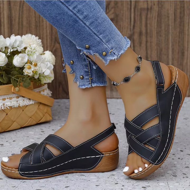 Damenfüße in schwarzen Sandalen mit Keilabsatz, kombiniert mit blauen Jeans. Im Hintergrund ein Korb mit weißen Blumen. Sommermode, bequeme Schuhe.