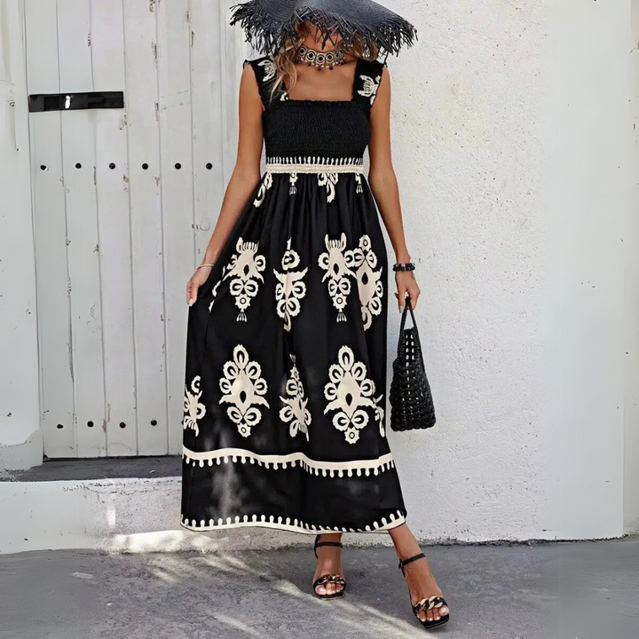 Frau in schwarzem, ärmellosem Maxikleid mit weißem Blumenmuster, trägt schwarzen Hut und Sandalen, steht vor weißer Wand. Sommermode, Boho-Stil.