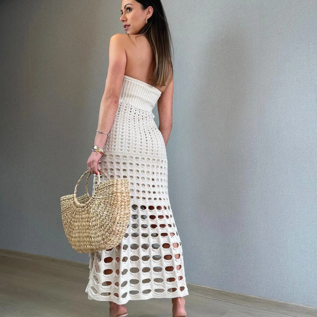 Frau in weißem, gehäkeltem Kleid mit Strohtasche, steht vor grauer Wand. Sommermode, elegantes Outfit, trendige Accessoires.