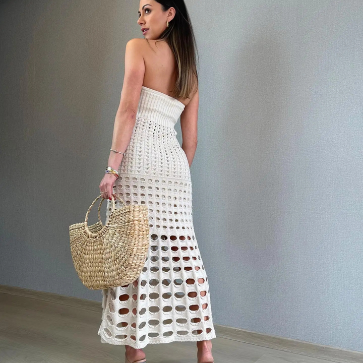 Frau in weißem, gehäkeltem Kleid mit Strohtasche, steht vor grauer Wand. Sommermode, elegantes Outfit, trendige Accessoires.