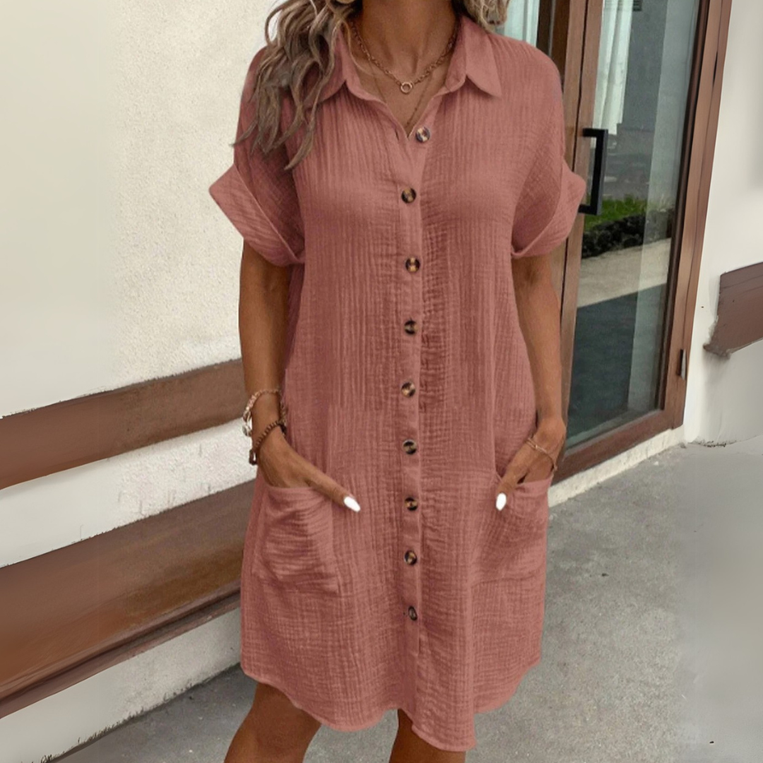 Frau in lockerem, knielangem, rosafarbenem Hemdkleid mit Knopfleiste und Taschen, steht vor modernem Gebäude. Sommermode, lässiger Stil.