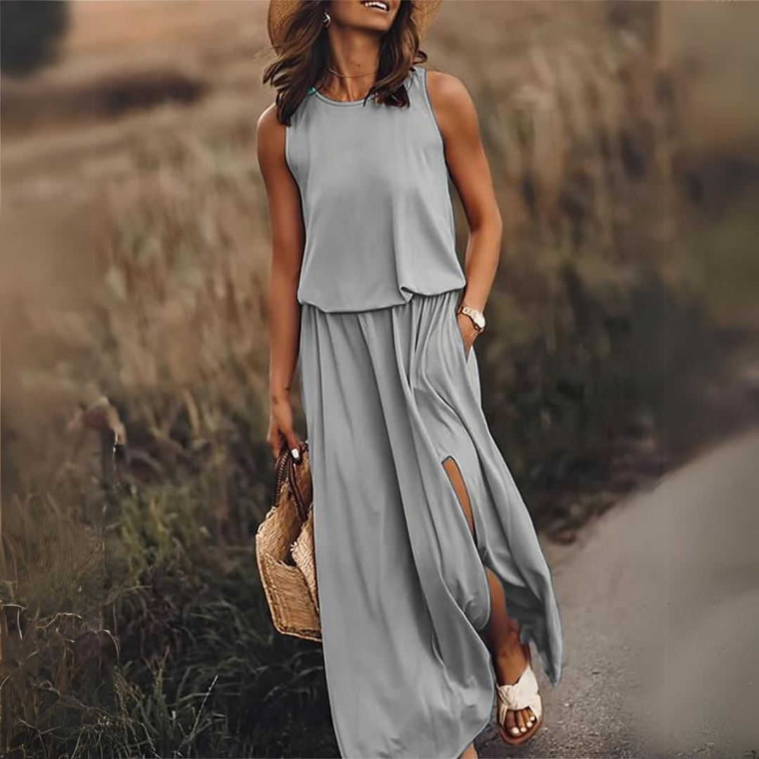 Frau in grauem Maxikleid mit Schlitz, Strohhut und Sandalen spaziert auf ländlichem Weg, trägt geflochtene Tasche. Sommermode, Freizeitkleidung.