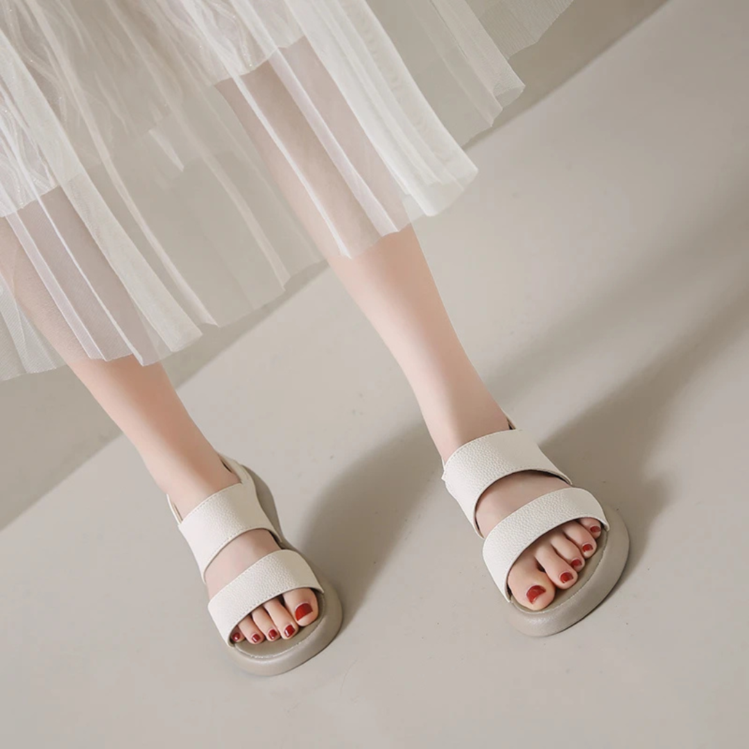 Weiße Sandalen mit breiten Riemen an Frauenfüßen, kombiniert mit einem weißen, transparenten Rock. Elegante Sommermode, minimalistisch und stilvoll.