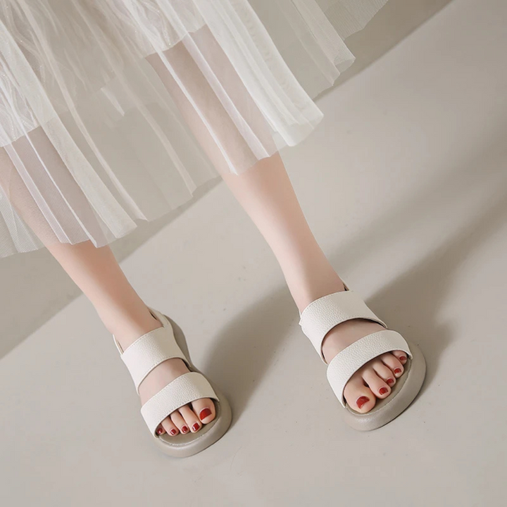 Weiße Sandalen mit breiten Riemen an Frauenfüßen, kombiniert mit einem weißen, transparenten Rock. Elegante Sommermode, minimalistisch und stilvoll.