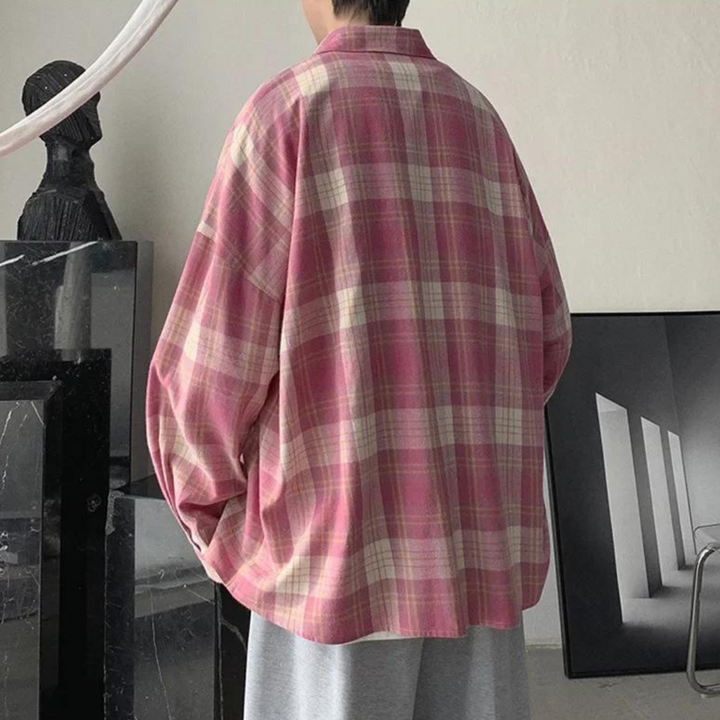 Person in übergroßem, rosa kariertem Hemd und grauer Hose steht in modernem Raum. Stilvolle, lässige Mode, minimalistische Inneneinrichtung.