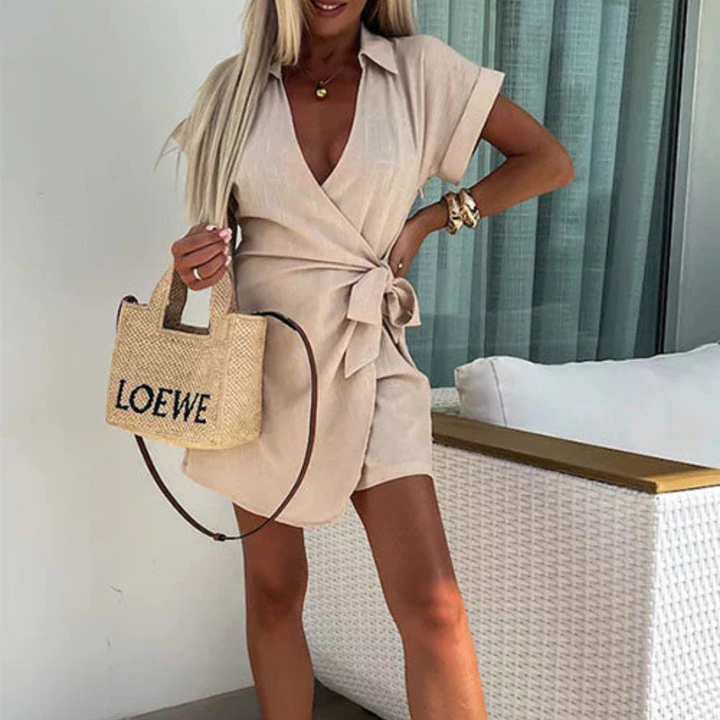 Frau in beigem Wickelkleid mit Strohtasche, steht vor einem modernen Sofa. Sommermode, eleganter Look, Freizeitkleidung, stilvolle Accessoires.