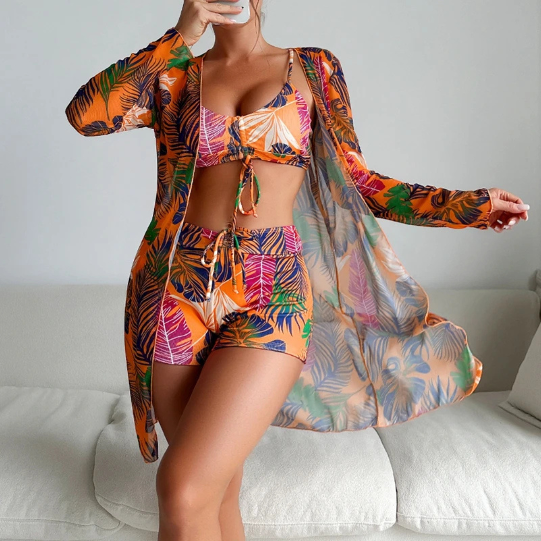 Frau in tropischem, buntem Strandoutfit mit Blättermuster, bestehend aus Bikini, Shorts und Kimono, posiert auf weißem Sofa. Sommermode, Strandkleidung.