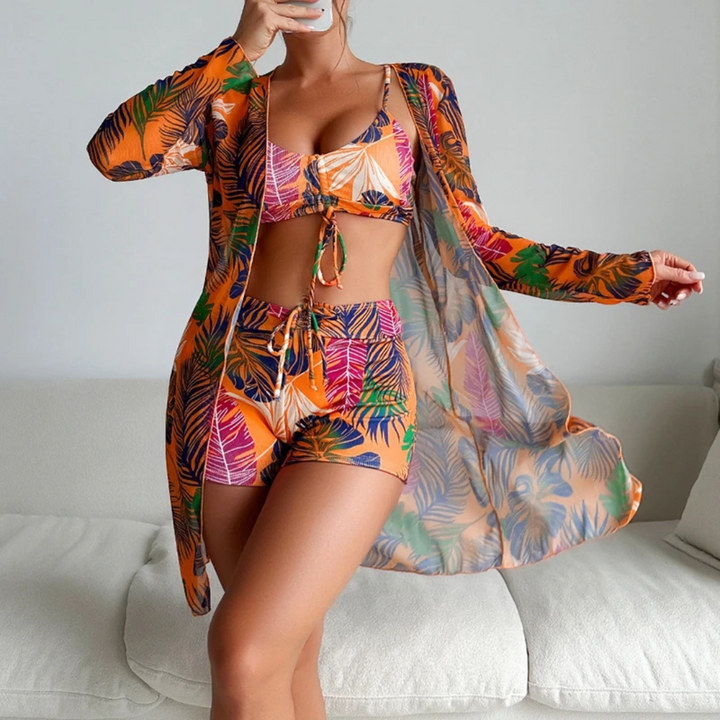 Frau in tropischem, buntem Strandoutfit mit Blättermuster, bestehend aus Bikini, Shorts und Kimono, posiert auf weißem Sofa. Sommermode, Strandkleidung.