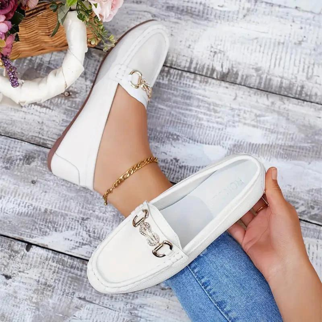 Weiße Damen-Loafer mit goldener Schnalle, getragen von einer Person in blauen Jeans, auf einem rustikalen Holzboden. Elegante Schuhe, modisches Design.