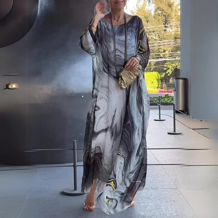 Frau in elegantem, langem Kleid mit abstraktem Muster und goldener Tasche, steht in modernem Innenraum. Mode, Damenbekleidung, stilvoll.