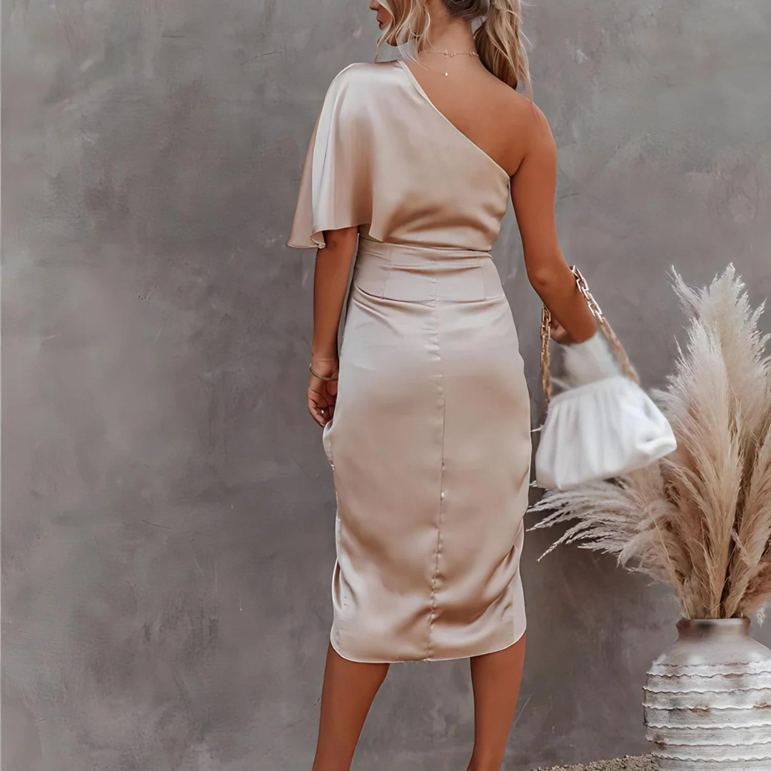 Frau in eleganten, einärmeligen, beigen Satin-Kleid, hält weiße Handtasche. Steht vor grauer Wand, neben Pampasgras. Fashion, Abendmode, Damenkleidung.