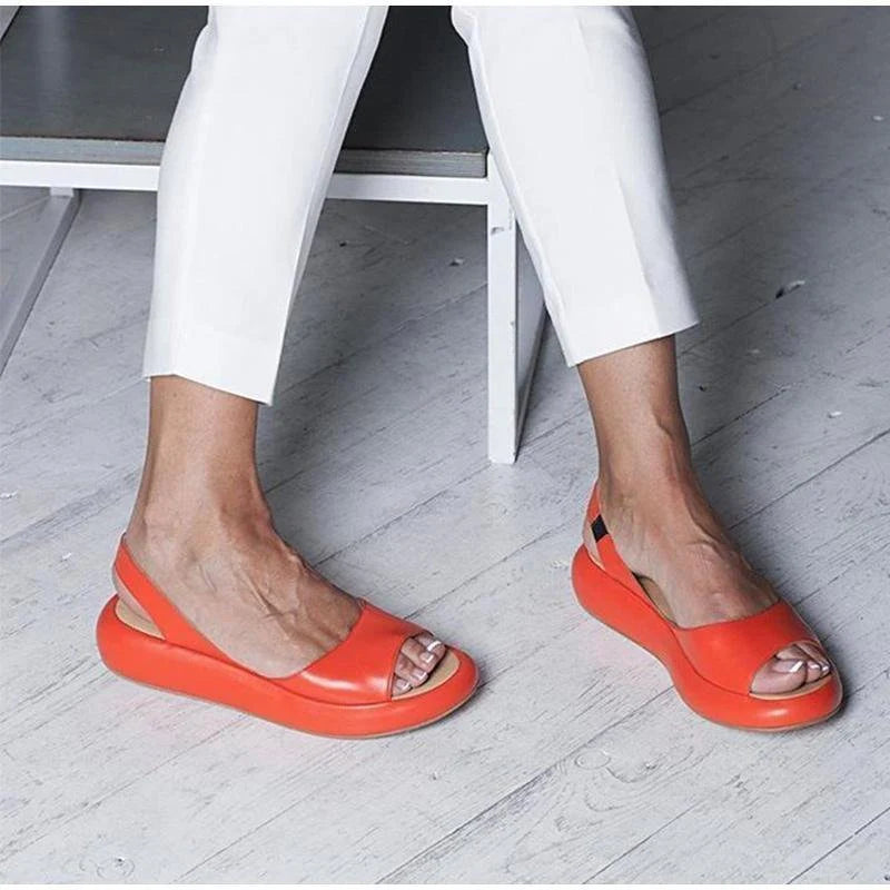 Rote Sandalen mit Plateauabsatz an Frauenfüßen, kombiniert mit weißen Hosen auf hellem Holzboden. Modische Sommerschuhe, Damenmode.