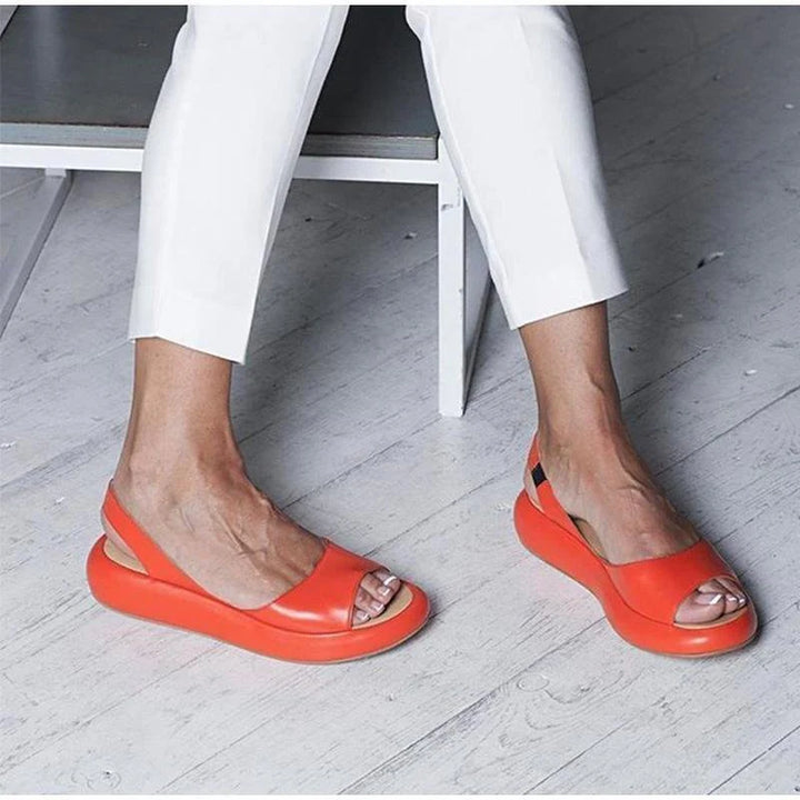 Rote Sandalen mit Plateauabsatz an Frauenfüßen, kombiniert mit weißen Hosen auf hellem Holzboden. Modische Sommerschuhe, Damenmode.