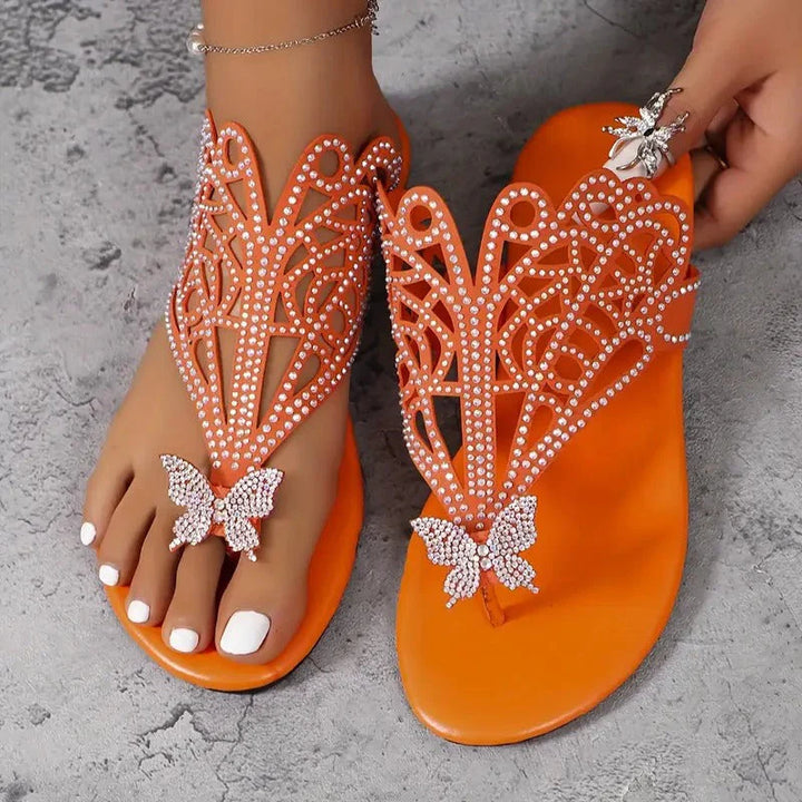 Orange Sandalen mit Schmetterlingsdesign und Strasssteinen, eleganter Sommerschuh auf grauem Hintergrund, modische Damenfußbekleidung.