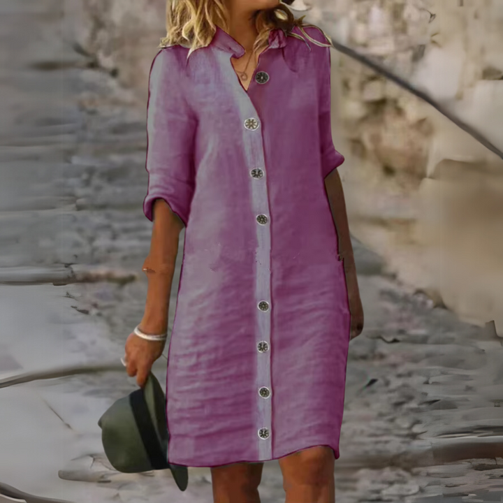 Frau in lila Leinenkleid mit Knöpfen, hält Hut, geht auf gepflastertem Weg. Sommermode, Freizeitkleidung, stilvoll, lässig.