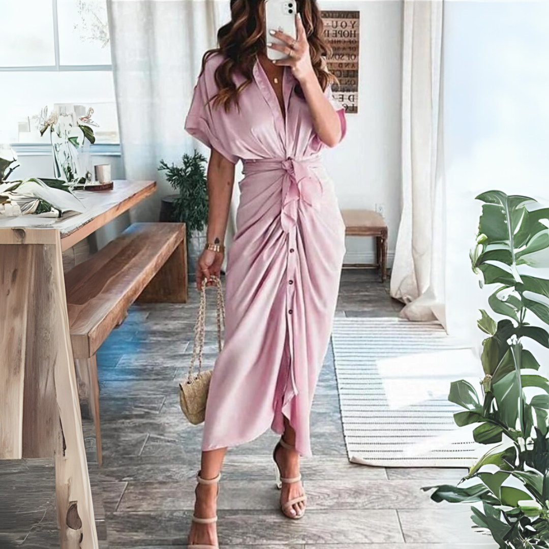 Frau in rosa Hemdkleid mit Taillenknoten, steht in modernem Wohnzimmer mit Holztisch und Pflanzen, hält eine kleine Handtasche.