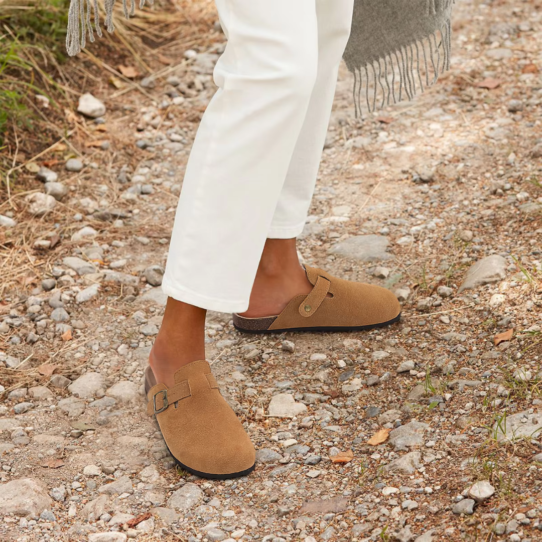 Braune Wildleder-Clogs auf einem steinigen Weg, getragen von einer Person in weißen Hosen. Bequeme, modische Schuhe für Outdoor-Aktivitäten.