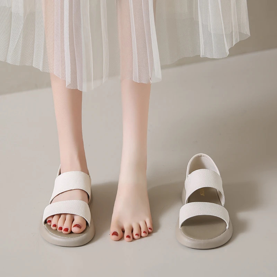Weiße Sandalen auf grauem Boden, neben nackten Füßen mit rotem Nagellack. Elegante Damenmode, Sommertrends, bequeme Schuhe.