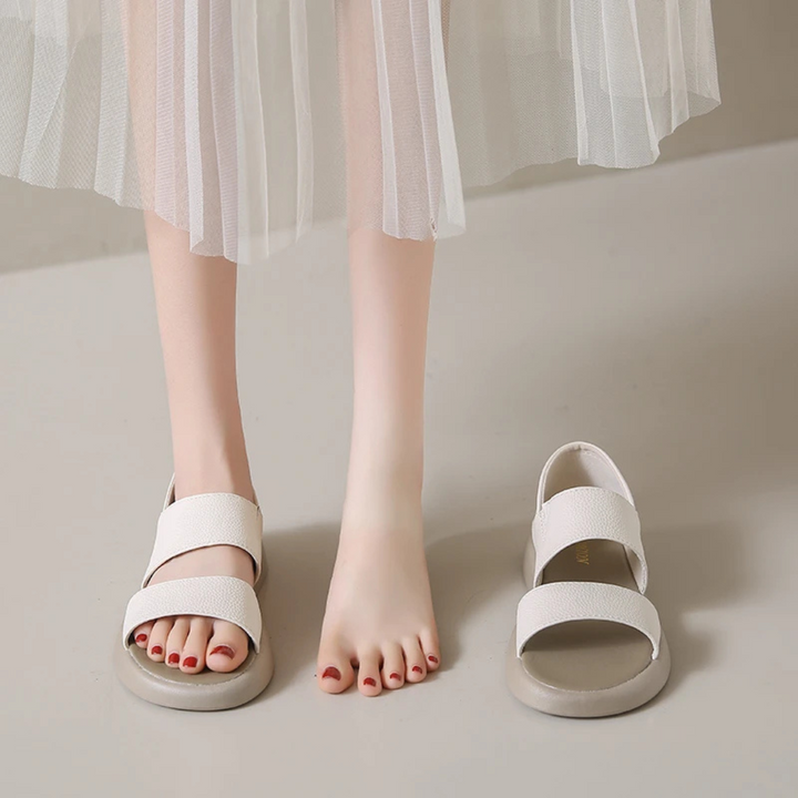 Weiße Sandalen auf grauem Boden, neben nackten Füßen mit rotem Nagellack. Elegante Damenmode, Sommertrends, bequeme Schuhe.