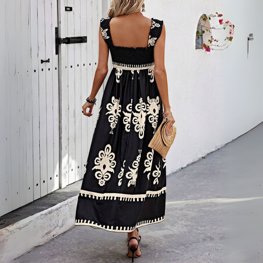 Frau in schwarzem Maxikleid mit weißem Blumenmuster, hält eine Strohtasche, geht auf einem gepflasterten Weg vor einer weißen Wand.