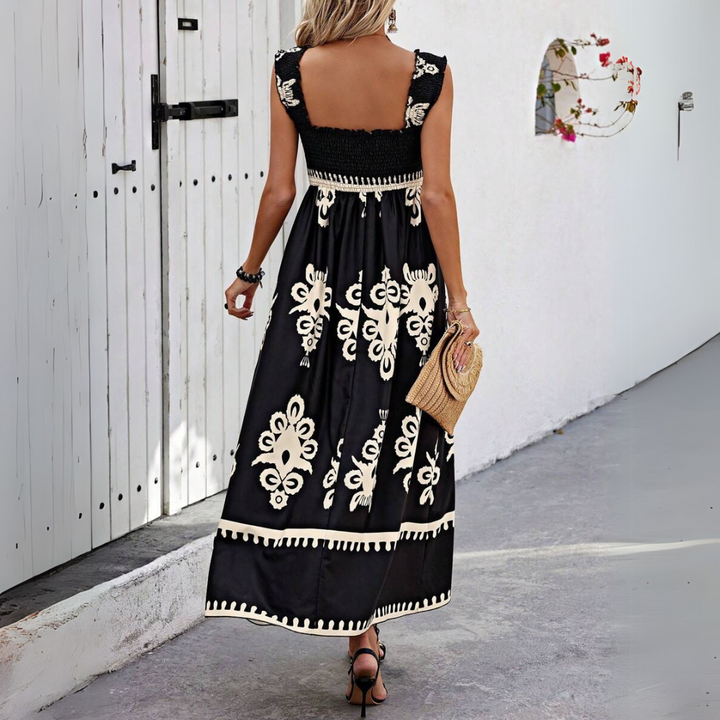 Frau in schwarzem Maxikleid mit weißem Blumenmuster, hält eine Strohtasche, geht auf einem gepflasterten Weg vor einer weißen Wand.