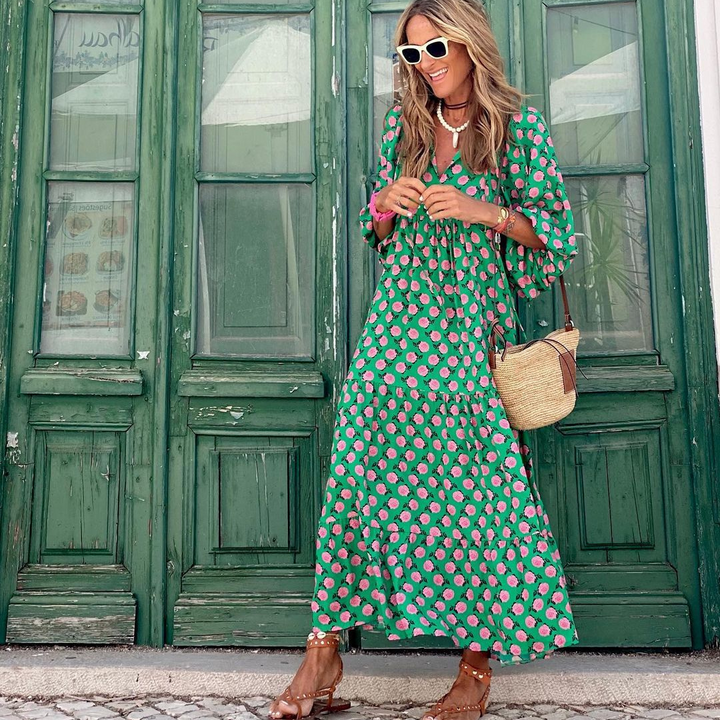 Frau in grünem, geblümtem Maxikleid vor grüner Holztür, trägt Sonnenbrille und Strohtasche. Sommermode, Boho-Stil, lässiger Look.