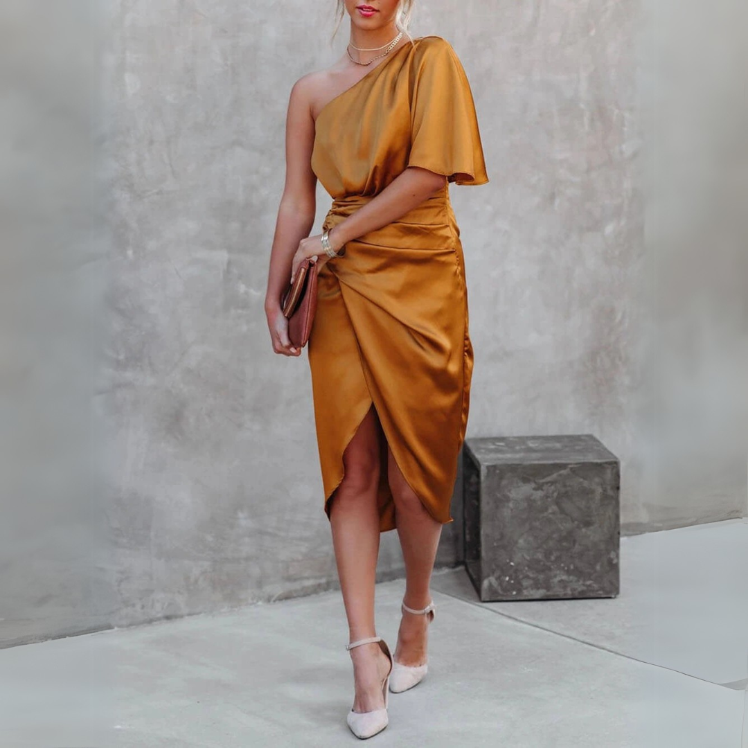 Frau in eleganten, einärmeligen, goldenen Satin-Kleid mit asymmetrischem Schnitt, hält eine Clutch, trägt weiße High Heels, steht vor grauer Wand.