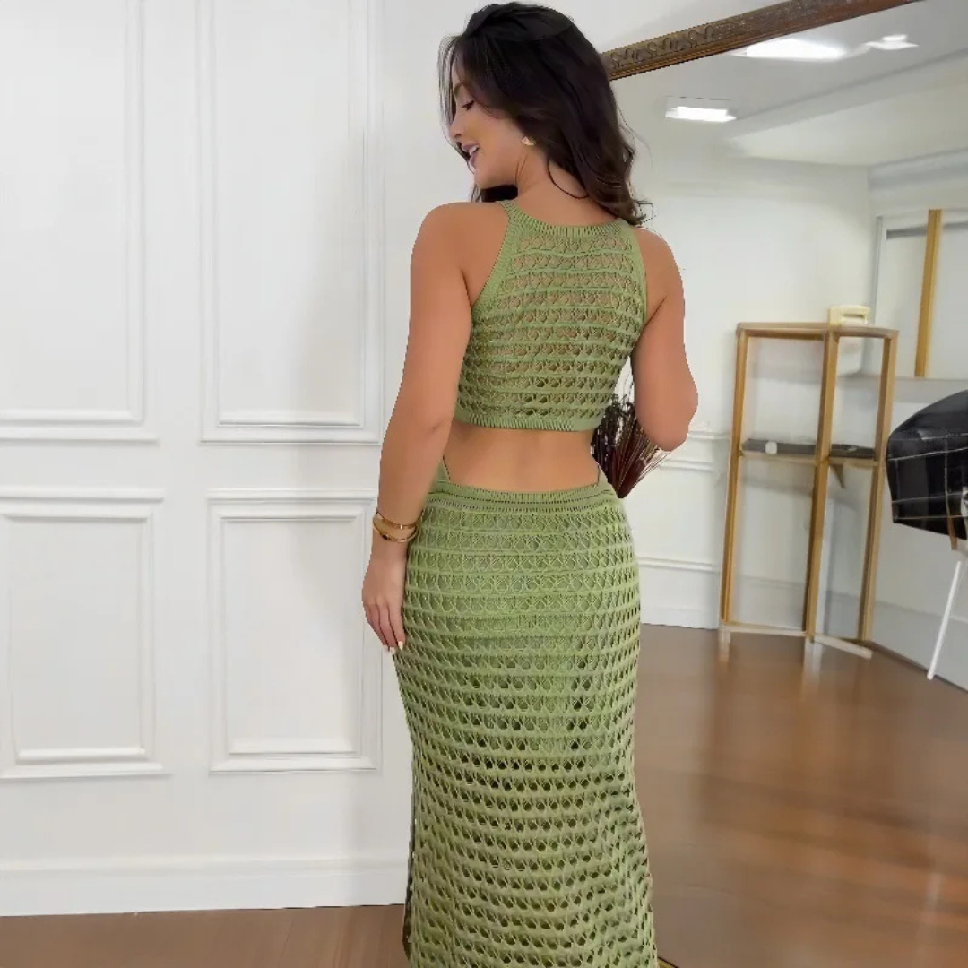 Frau in grünem, gehäkeltem Zweiteiler-Kleid, steht in modernem Raum mit Holzfußboden und Spiegel. Elegante Mode, Sommeroutfit, Häkeltrend.