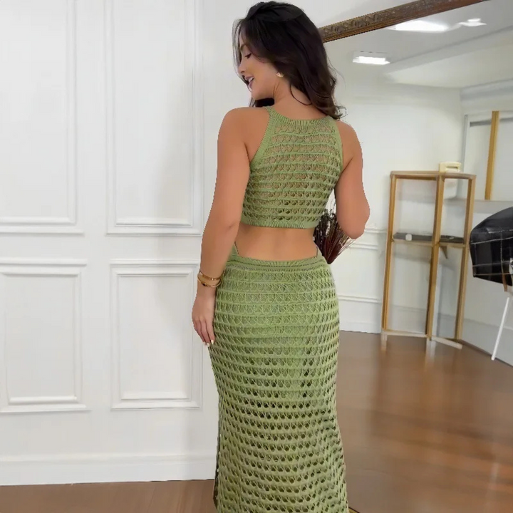 Frau in grünem, gehäkeltem Zweiteiler-Kleid, steht in modernem Raum mit Holzfußboden und Spiegel. Elegante Mode, Sommeroutfit, Häkeltrend.