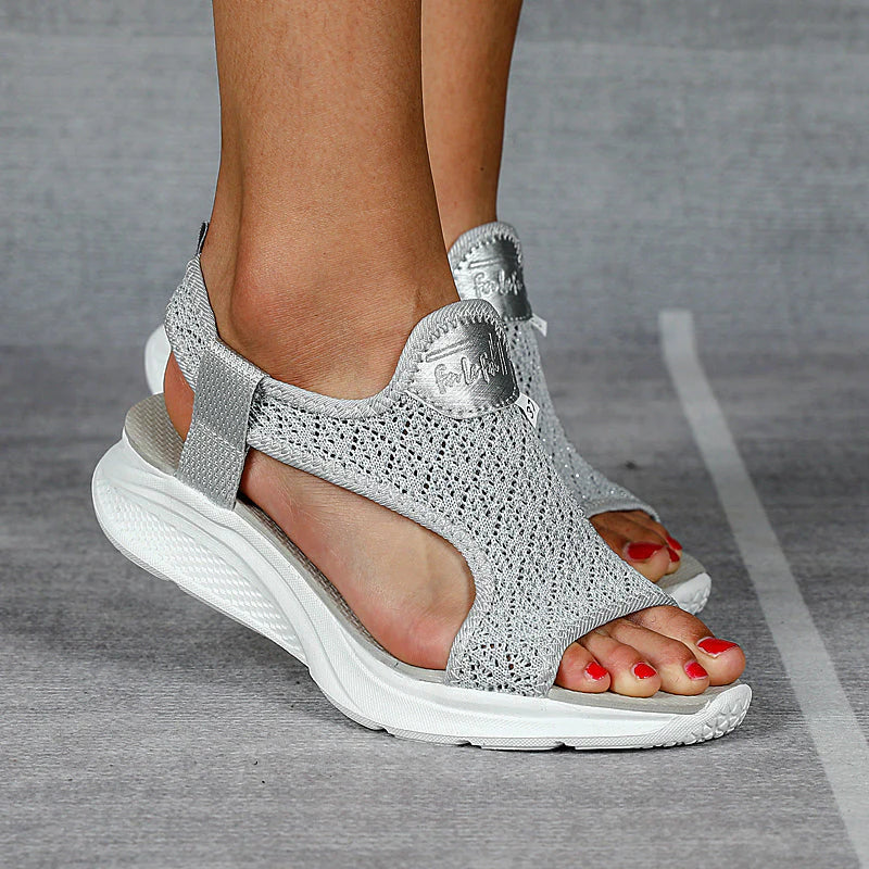 Damenfüße in silbernen, offenen Sandalen mit Keilabsatz und Strickmuster auf grauem Hintergrund. Bequeme Sommerschuhe, modisch und elegant.