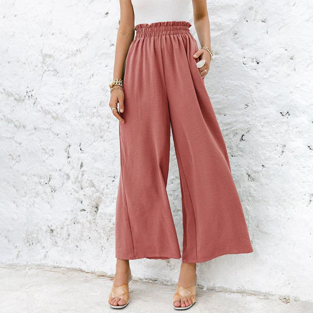 Frau in weiten, rosafarbenen Palazzo-Hosen vor weißer Wand, trägt Sandalen und Armbänder. Modetrend, Sommeroutfit, Damenbekleidung.