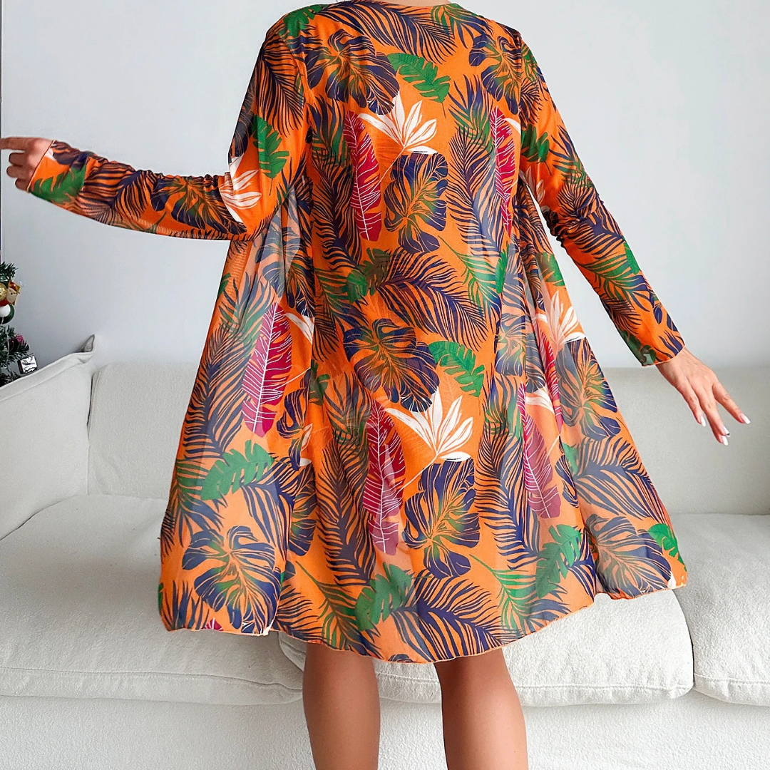 Frau in orangefarbenem Kleid mit tropischem Blattmuster, steht auf weißem Sofa. Modetrend, tropisches Design, farbenfrohes Kleid, Inneneinrichtung.