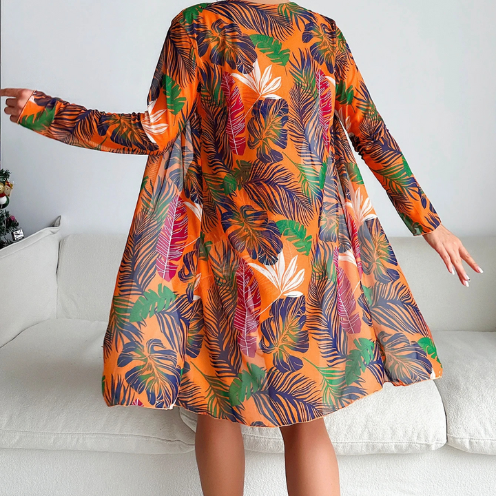 Frau in orangefarbenem Kleid mit tropischem Blattmuster, steht auf weißem Sofa. Modetrend, tropisches Design, farbenfrohes Kleid, Inneneinrichtung.