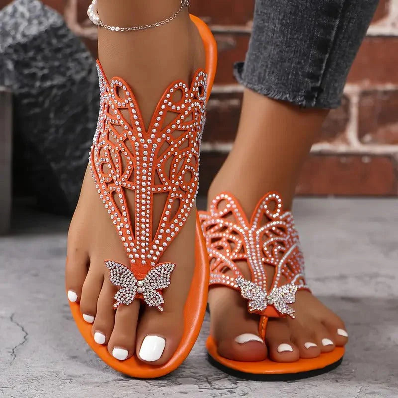 Orange Sandalen mit Strassverzierung und Schmetterlingsdesign, getragen von einer Person mit weiß lackierten Zehennägeln, vor einer Ziegelwand.
