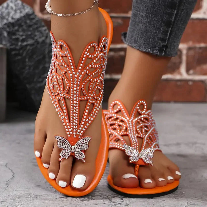 Orange Sandalen mit Strassverzierung und Schmetterlingsdesign, getragen von einer Person mit weiß lackierten Zehennägeln, vor einer Ziegelwand.