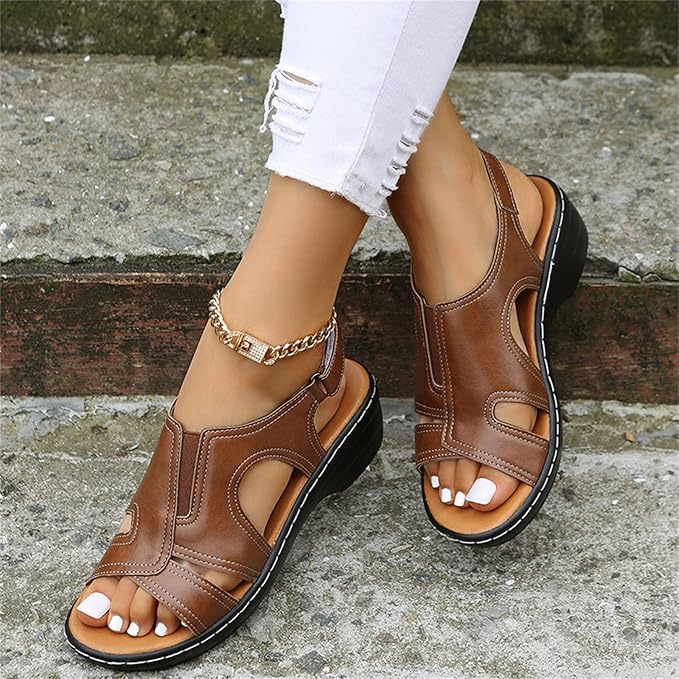 Damenfüße in braunen Ledersandalen mit Keilabsatz und goldener Fußkette, getragen auf einer Steintreppe. Perfekt für Sommermode und Komfort.