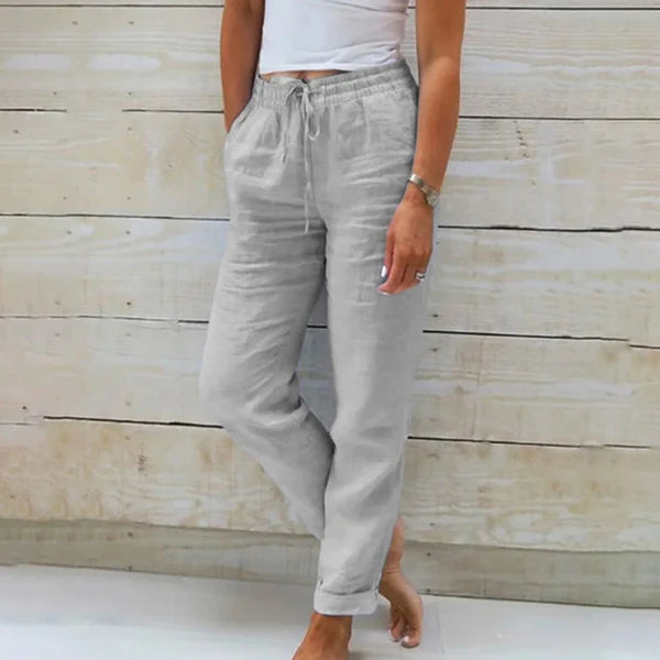 Frau in grauer Leinenhose und weißem Top vor Holzwand. Bequeme Sommermode, lässiger Stil, Damenbekleidung, Freizeitlook.