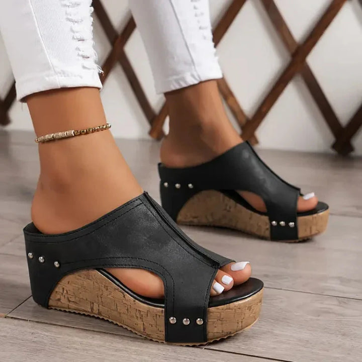 Schwarze Keilsandaletten mit Korksohle und Nieten, getragen von einer Person in weißen Jeans. Modische Sommerschuhe, ideal für Damen.