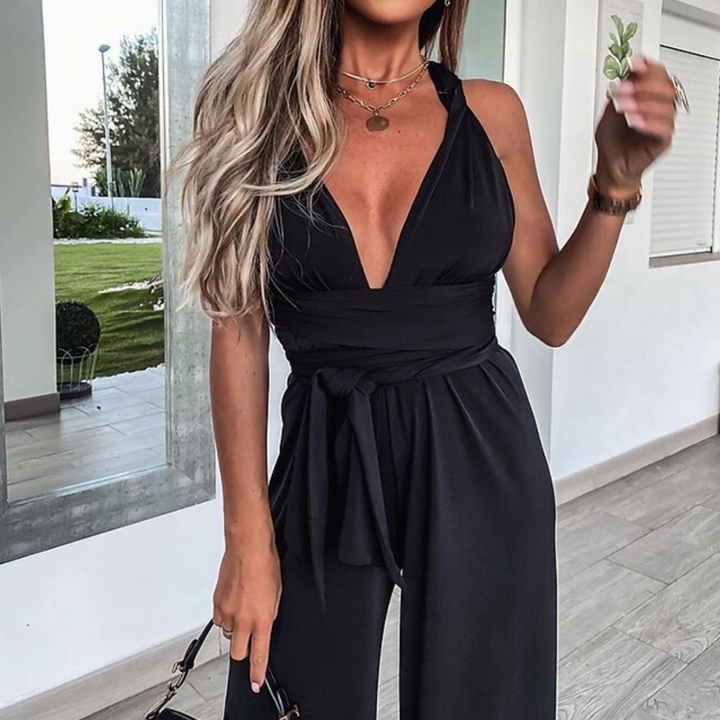 Frau in elegantem schwarzen Jumpsuit mit tiefem V-Ausschnitt, modischen Accessoires und blonden Haaren, steht in modernem Innenraum.