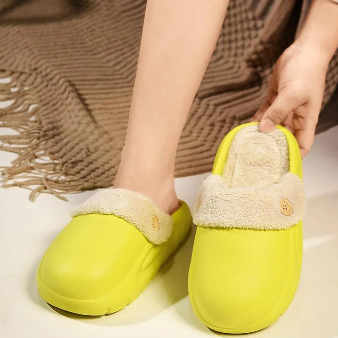 Gelbe Hausschuhe mit weichem, beigem Futter und Knopfdetail, getragen auf einem Teppich. Bequeme, warme Pantoffeln für Zuhause.