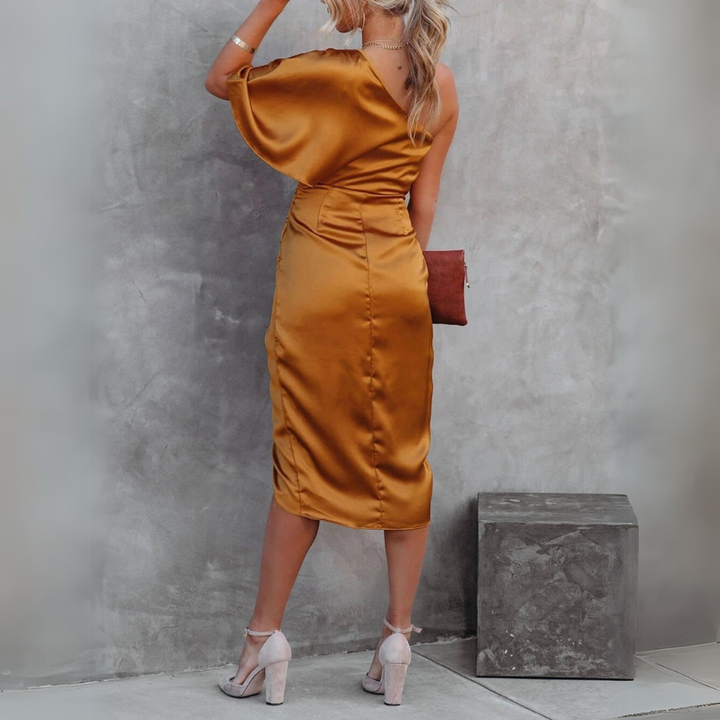 Frau in elegantem, goldfarbenem, schulterfreiem Satinkleid, steht vor grauer Wand, hält braune Clutch, trägt hohe Absätze. Fashion, Abendmode.