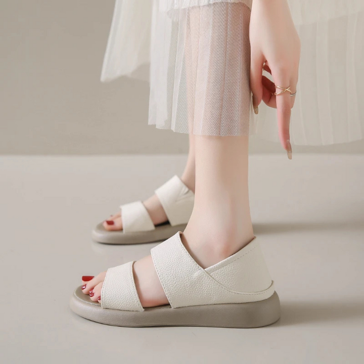 Weiße Sandalen mit breiten Riemen an Frauenfüßen, kombiniert mit einem transparenten, weißen Rock. Modische Sommerschuhe, bequem und stilvoll.