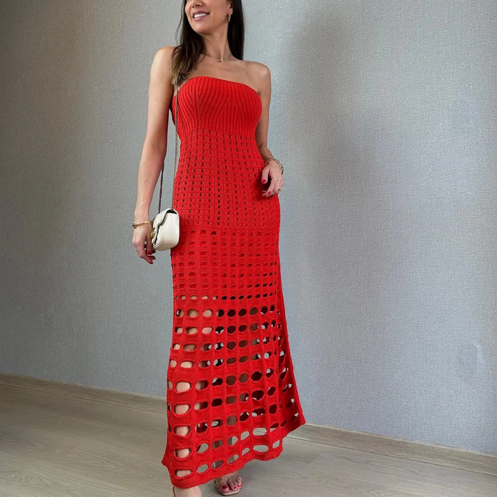 Frau in rotem, schulterfreiem Strickkleid mit Lochmuster, steht vor grauer Wand. Elegantes Abendoutfit, modisch, stilvoll, mit weißer Handtasche.