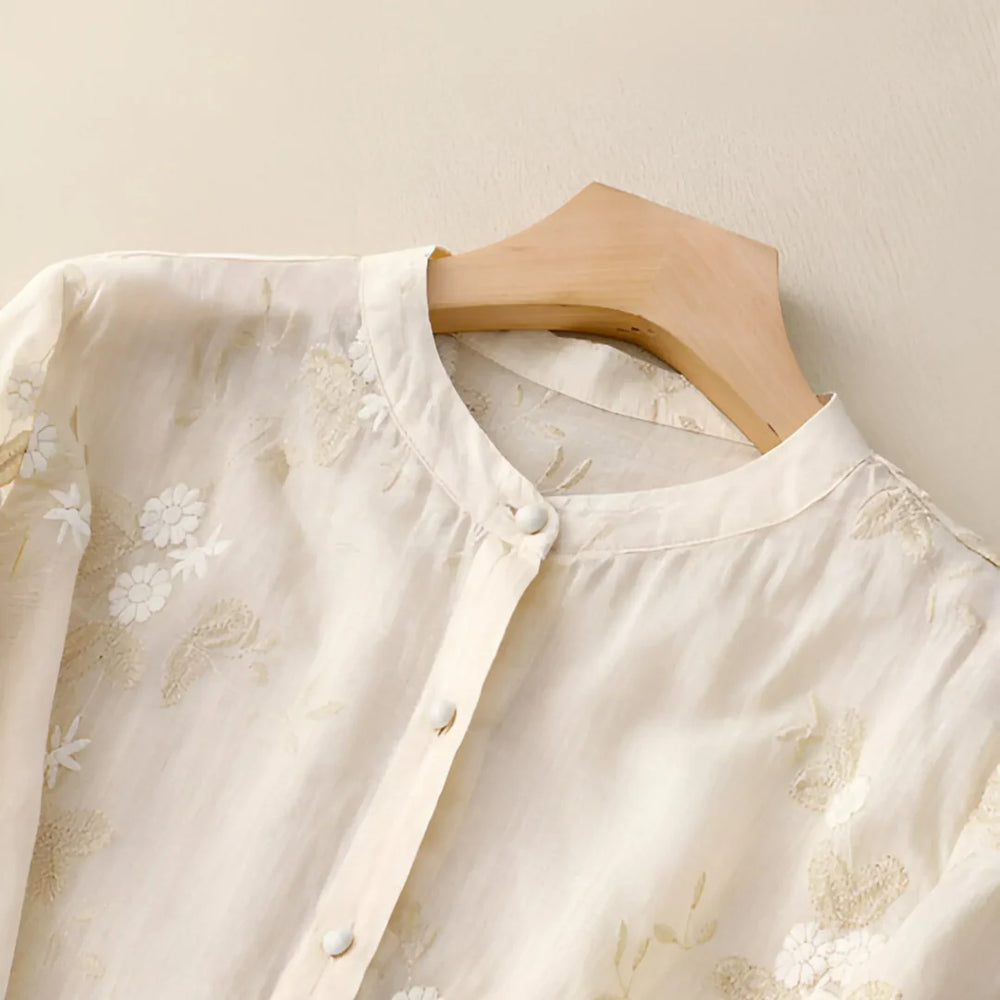 Elegante cremefarbene Bluse mit floralen Stickereien, Stehkragen und Knopfleiste auf Holzkleiderbügel. Damenmode, Frühlingskollektion, stilvoll.