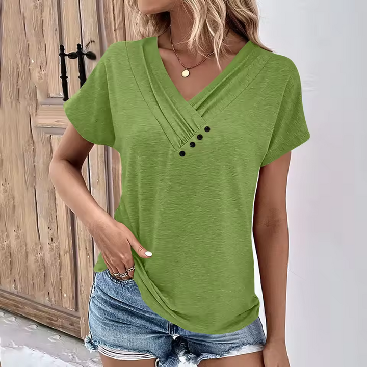 Frau in grünem V-Ausschnitt-T-Shirt mit Knöpfen, kombiniert mit blauen Jeansshorts, steht vor einer Holztür. Sommermode, lässiger Stil.
