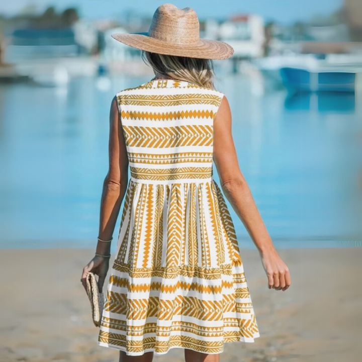 Frau in Sommerkleid mit geometrischem Muster und Strohhut am Strand, hält eine Tasche. Hintergrund: Meer und Boote. Ideal für Sommermode und Strandurlaub.